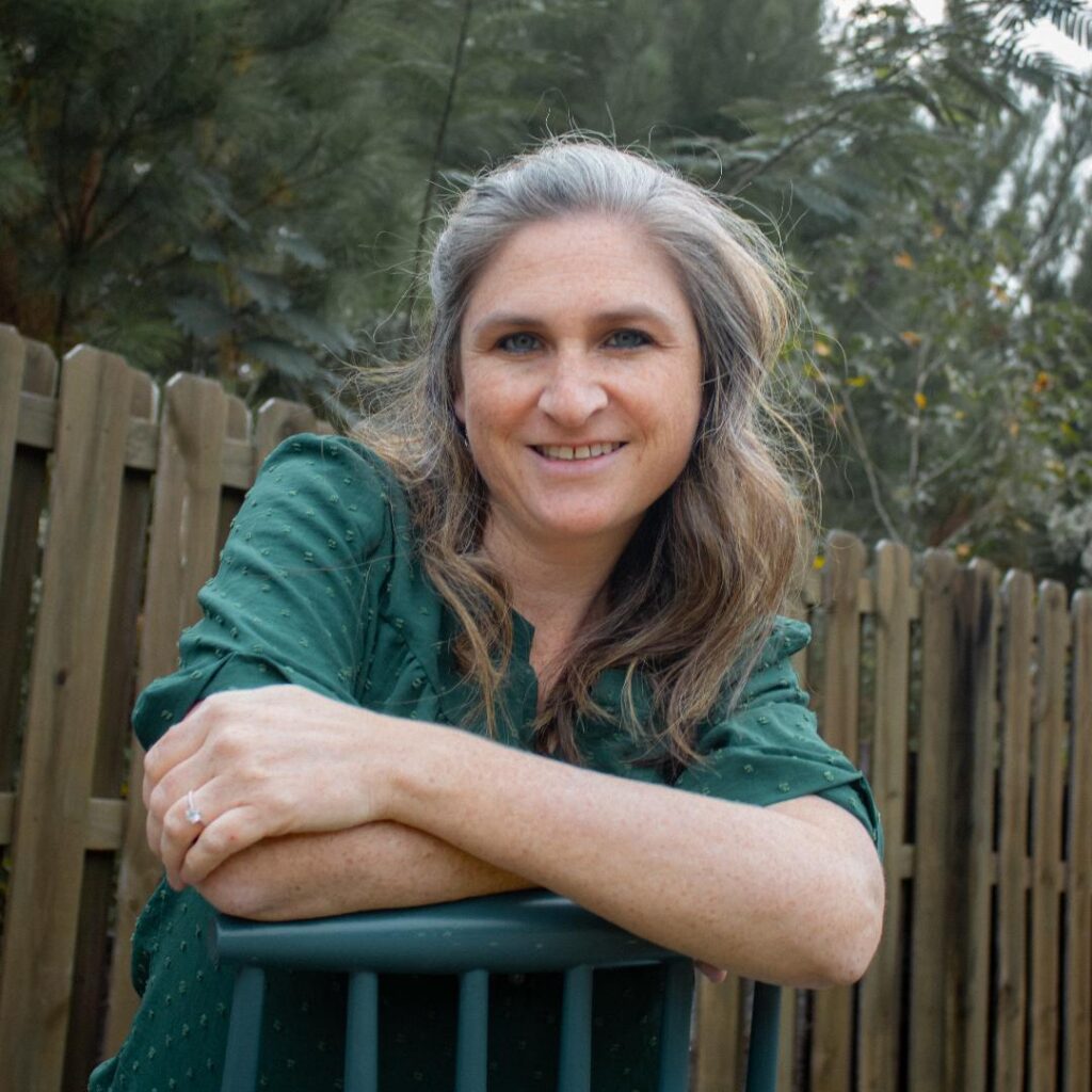 Melissa Burch is a Caucasian woman with long gray/brown hair. She is wearing a green button down shirt leaning on a railing.