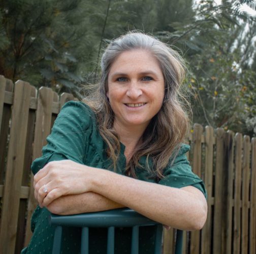 Melissa Burch is a Caucasian woman with long gray/brown hair. She is wearing a green button down shirt leaning on a railing.