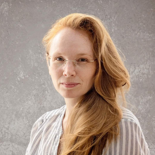 Eva Creel is a Caucasian woman with long, strawberry blonde hair. She is wearing glasses and a stripped blouse,
